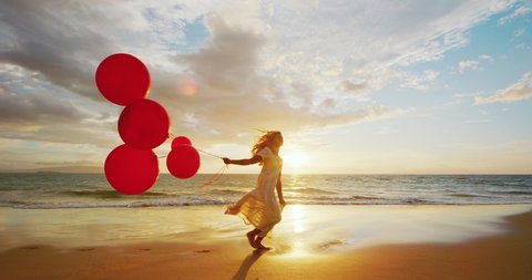 Happy young woman with red balloons running down the beach at sunset, freedom and happiness concept