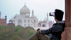 A young man sitting and clicking a picture of Taj Mahal Mausoleum using a smartphone camera while enjoying the beautiful view. A male tourist or traveler on holidays taking a photo on a mobile phone 