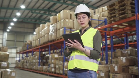 Young chinese warehouse operator woman in safety hat working on digital tablet while standing in large factory stockroom.