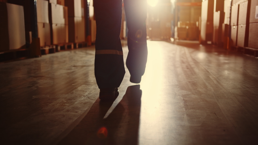 Professional Warehouse Worker Wearing Safety West and Hard Hat Walking Through The Strorage Facility, Distribution, Delivery Center. Emotional, Moody Following Shot Starting with Elevating Low Angle Royalty-Free Stock Footage #1060977049