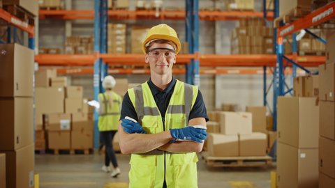 Handsome Smiling Worker Wearing Hard Hat, Crossing Arms Standing in the Retail Warehouse full of Shelves with Cardboard Boxes. Professional Working in Logistics, Delivery, and Distribution Center
