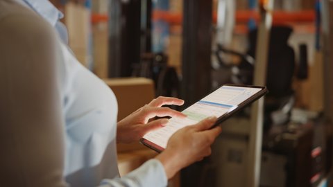 In Warehouse Manager Uses Digital Tablet with App to Check Package Delivery Status with Graphs, Infographics, and Statistics on Screen. Distribution Center with Shelves with Cardboard Boxes