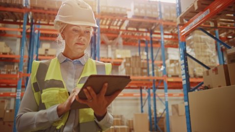 Professional Confident Worker Wearing Hard Hat Checks Stock and Inventory with Digital Tablet Computer in the Retail Warehouse full of Shelves with Goods. Working in Logistics, Distribution Center
