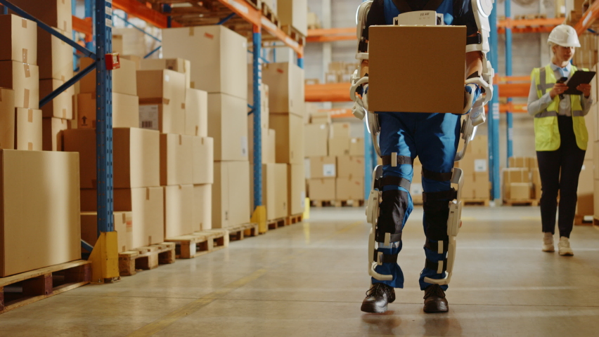 High-Tech Futuristic Warehouse: Worker Wearing Advanced Full Body Powered Exoskeleton, Walks with Heavy Cardboard Box. Delivery Exosuit amplifies Human Strenght. Elevating Dolly Slow Motion Shot Royalty-Free Stock Footage #1060977160