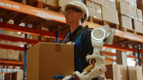 High-Tech Futuristic Warehouse: Worker Wearing Advanced Full Body Powered Exoskeleton, Walks with Heavy Cardboard Box. Delivery Exosuit amplifies Human Strenght. Elevating Dolly Slow Motion Shotの動画素材