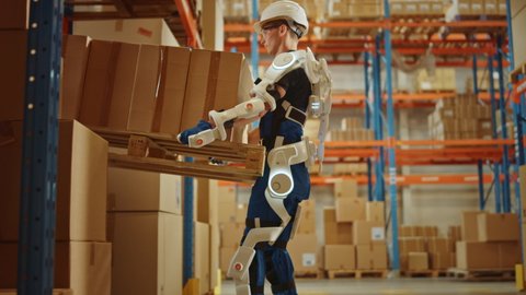 High-Tech Futuristic Warehouse: Worker Wearing Advanced Full Body Powered exoskeleton, Lifts and Walks with Heavy Pallet full of Cardboard Boxes. Delivery Exosuit amplifies Strenght. Following Shot