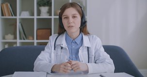 female retiree doctor is consulting online, looking at camera and nodding, listening by headphones, telemedicine service