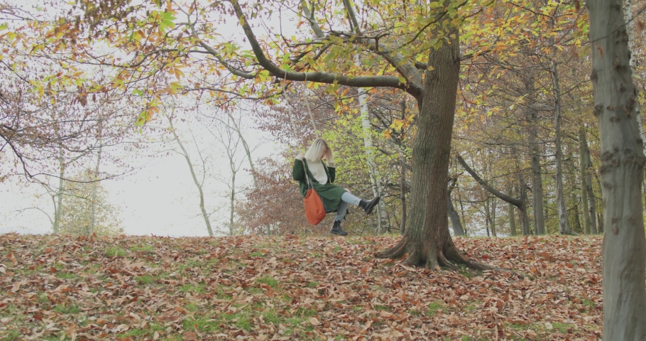 Slow motion approach of a young and lonely woman sitting on a swing under a big tree. Young woman going back to being a child.
