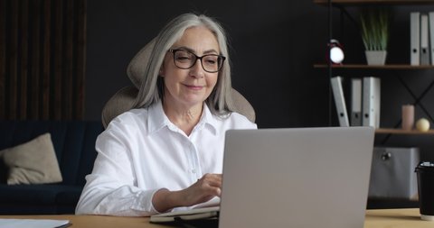 Attractive old Female Lecturer working Indoor on Laptop. Old Mature Senior Woman Teacher, Lecturer, Coach holding a Seminar, Conference, Webinar. Education Indoor. Distantly Study.