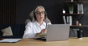 Old Mature Senior Woman Teacher, Lecturer, Coach with Headphones with Microphone, holding a Seminar, Conference, Webinar. Progressive old Female Lecturer working Indoor on Quarantine. Education.