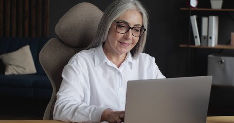 Portrait of Happy mature middle-aged Businesswoman smiling, texting Messages on Laptop. Attractive old senior Female  working on Freelance from Home Office, Progressive People, World. Technology.