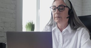 Close Up of mature senior Woman working indoor, as Operator in Call-Center, Technical Support. Confident Businesswoman talking speaking in Headset, using Laptop. Easy Technology. Progress.
