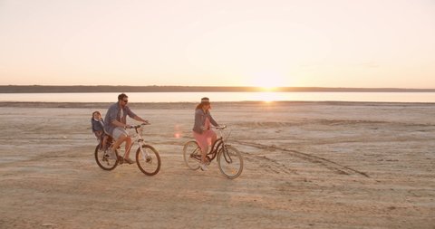 Family of three - mother, father and little son riding bicycles near the lake during beautiful sunset. Summer vacations concept.