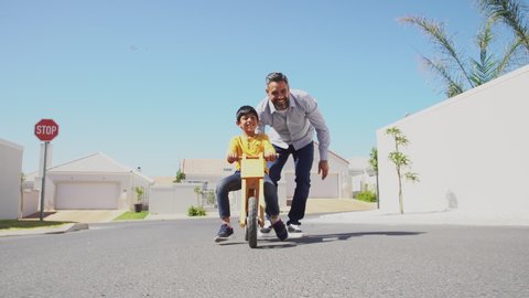Happy latin father helping smiling boy to ride wooden balance cycle on street. Happy middle eastern child and young dad riding bike in lane. Smiling daddy teaching son to ride a balance bicycle.