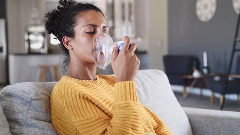 Black woman holding a mask nebulizer inhaling fumes medication into lungs. African sick lady inhaling through inhaler mask sitting on the couch with copy space. Self treatment of the respiratory tract