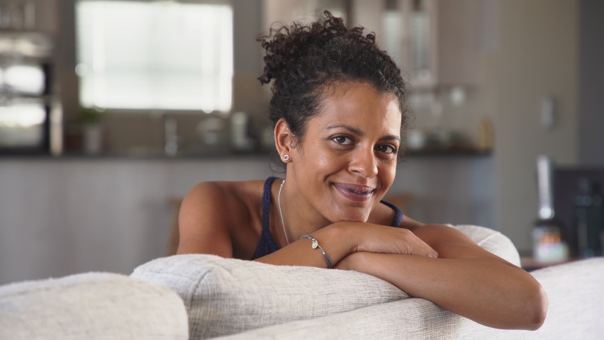 Cheerful ethnic mid adult woman relaxing on sofa at home. Successful young african woman with toothy smile sitting on couch and looking at camera. Portrait of confident black woman laughing. | Shutterstock HD Video #1060982692