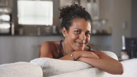 Cheerful ethnic mid adult woman relaxing on sofa at home. Successful young african woman with toothy smile sitting on couch and looking at camera. Portrait of confident black woman laughing.