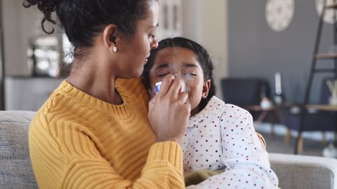 Black mother helping sick daughter use nebulizer while embracing her on couch at home. Woman makes inhalation with equipment to indian girl. Ill child lying on couch having respiratory illness helped 