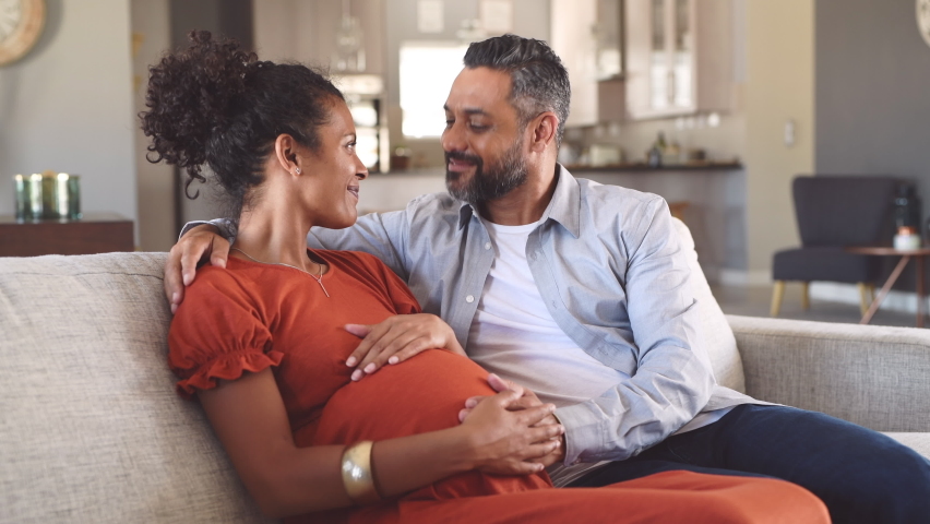 Mid adult couple expecting a baby while husband caresses the belly of his pregnant african woman. Happy father hands on expecting mother's baby bump while embracing her on couch. | Shutterstock HD Video #1060982704