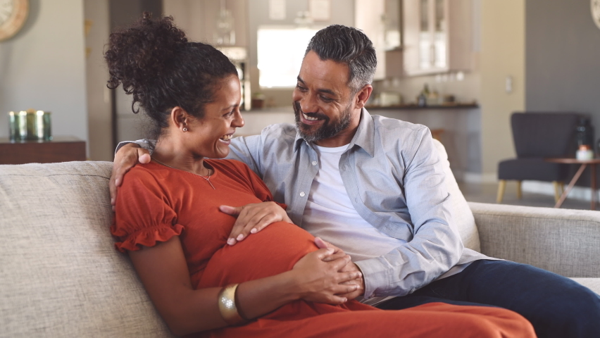 Mid adult couple expecting a baby while husband caresses the belly of his pregnant african woman. Happy father hands on expecting mother's baby bump while embracing her on couch.