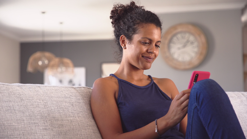 Cheerful young african woman using smartphone while sitting on couch. Black smiling woman using app on cellphone at home. Beautiful girl relaxing while chatting on mobile phone and looking at camera.  | Shutterstock HD Video #1060982707