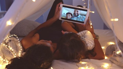 Mother and cute girl using digital tablet while lying in kid tent doing a video call with father and son. Indian family in online conversation with each other during quarantine and social distancing.