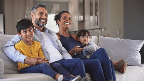 Happy indian man and african woman relaxing with daughter and son watching television at home together. Cheerful ethnic family relaxing on sofa at home watching movie with children.