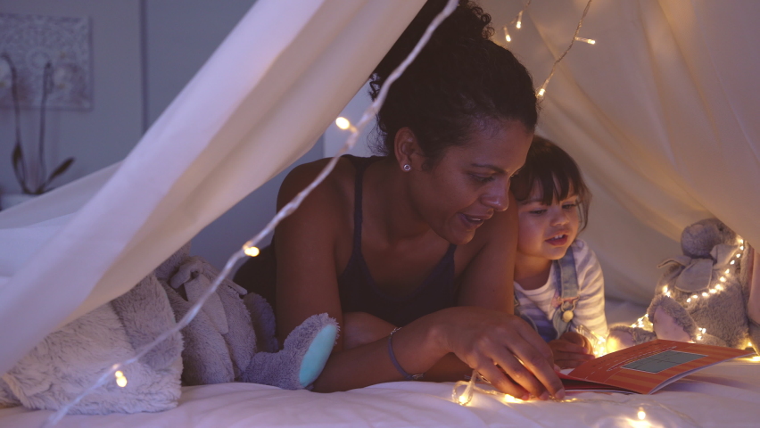 African american mother lying on bed in illuminated tent and playing with cute girl. Close up of mom telling a fairy tale in kid tent. Mother reading a story to little daughter before going to bed. Royalty-Free Stock Footage #1060982716