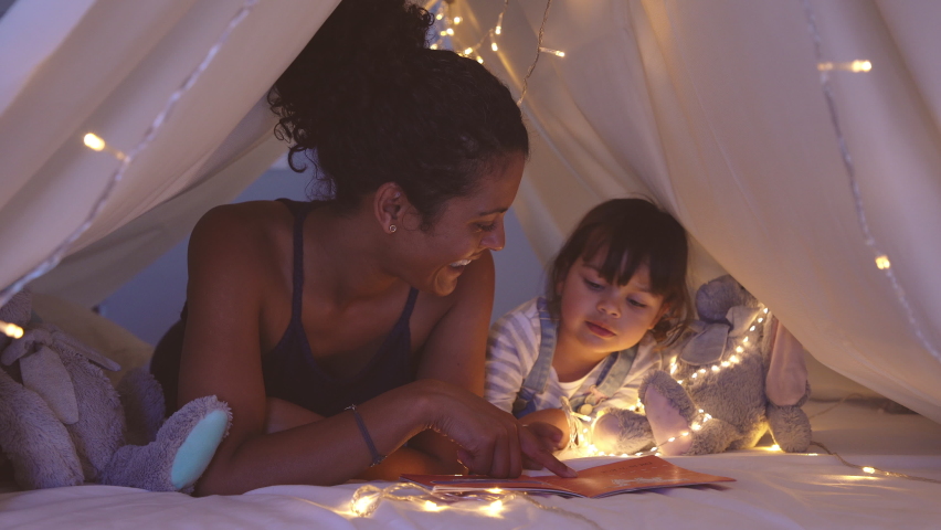 African american mother lying on bed in illuminated tent and playing with cute girl. Close up of mom telling a fairy tale in kid tent. Mother reading a story to little daughter before going to bed. | Shutterstock HD Video #1060982716
