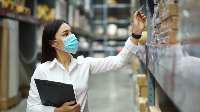 Woman worker with medical mask holding clipboard and checking inventory in the warehouse during coronavirus (covid-19) pandemic. | Shutterstock HD Video #1060982719