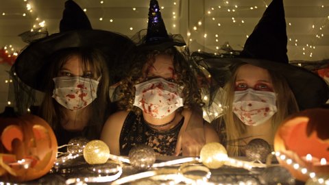 Three teenage girls in black costumes witchs with medical masks on their faces are sitting at a table. Halloween attributes on the table. Halloween during the covid19 coronavirus pandemic.