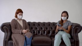 Two women wearing medical face mask, one shows the other something funny on the phone. Females sitting on a sofa.