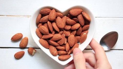 A heart shaped dish Full of almonds On to a white piece of wood