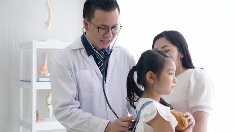 Asian pediatrician and kid concept. Middle-aged male doctor puts the stethoscope on the back of cute worried female child patient who is holding a teddy bear with the mother standing beside.