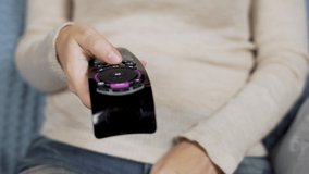 Woman Turns on the TV by Pressing the Button on the Remote Control from the TV Sitting on the Sofa, Front View. Female Fingers and TV Remote Control, Close-up.