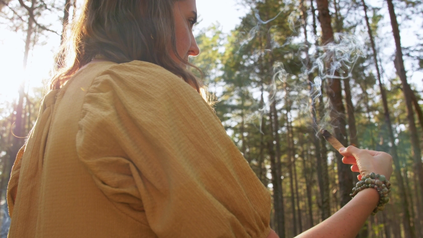 occult science and supernatural concept - young woman or witch with smoking palo santo stick performing magic ritual in forest