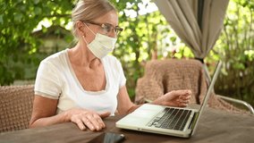 Technology, old age people concept - elderly senior woman takes off protective face mask working online with laptop computer outdoor in the garden. Remote work, distance education. 