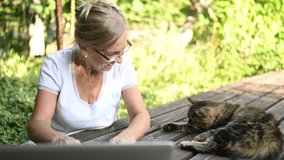 Technology, old age people concept - elderly happy senior woman with home cat working online with laptop computer outdoor in the garden. Remote work, distance education. 