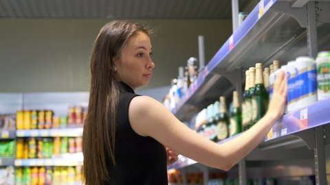 Young customer, woman is choosing beer in alcohol department of grocery store. She is taking a can from the rack, reading label and composition. Close up, slow motion
