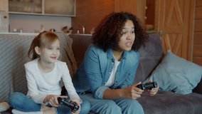 Slow motion of cute girl playing video game with Afro-American nanny using joysticks having fun pushing each other laughing on couch at home