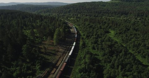Freight long train carries with cargo carriages in wild mountains landscape through a difficult part of Trans Siberian railways. Aerial drone wide view at summer day.