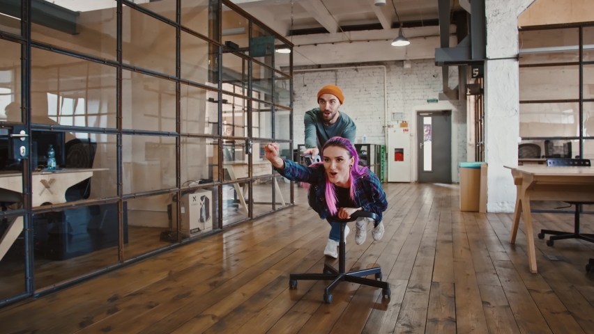 Flying to aim. Young woman lying on office chair, her coworker riding her in office, slow motion Royalty-Free Stock Footage #1061016265