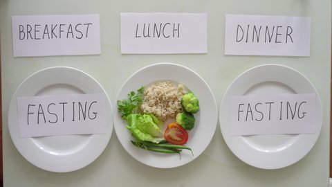 Hands of a woman put a plate with brown rice and vegetables under the title lunch, and under the title breakfast and dinner she puts on empty plates titles fasting. Interval fasting concept. Skipping