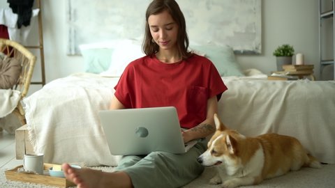 Young woman working with laptop remotely and spending day with pet in apartment interior.. Front view of beautiful young woman looking at computer screen and talking to dog while sitting on floor