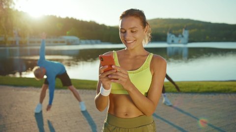 Joyful caucasian fitness trainer woman using mobile phone social media smiling while her friends training physical endurance doing gym exercises near lake background.