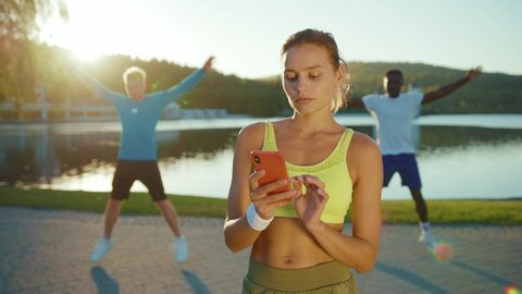 Beautiful muscular bodybuilder young woman using smartphone internet connecting with friends. Two athletic men doing cardio workout exercises at lake. Teamwork.
