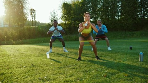 Mixed race handsome young people in team performing squats with elastic bands on the grass. Athletes training in public park beautiful nature on early morning. Sports.