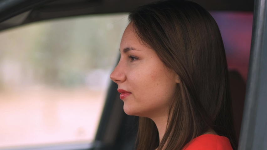 The beautiful young driver girl with red lipstick in a red dress turns and looks directly at the camera. The woman smiles. Red car