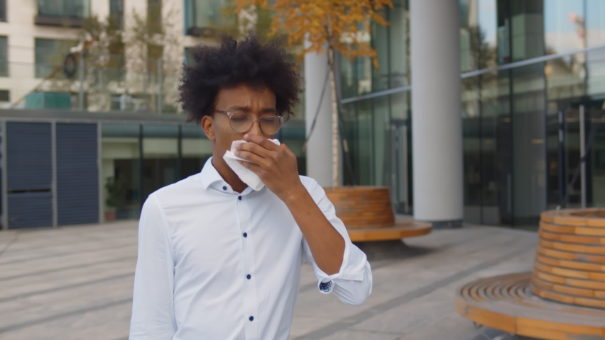 Young african businessman walking outside office building and sneezing in white napkin. Portrait of sick afro entrepreneur coughing in tissue walking outdoors business center | Shutterstock HD Video #1061025790