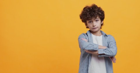 Portrait of little boy with folded arms expressing contempt or annoyance, looking at camera with condemnation, orange studio background